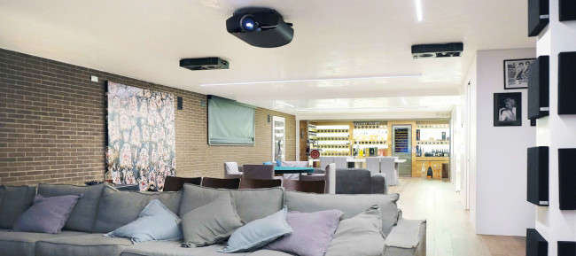 Una vista panoramica, dallo schermo, della grande sala dedicata allâ€™intrattenimento; in primo piano il bel proiettore Sony VPL-1100 mentre dietro a questo, installati a controsoffitto, si notano due  dei quattro diffusori surround. Sullo sfondo la bella cantina;   a destra in basso, una zona antistante alla sala.